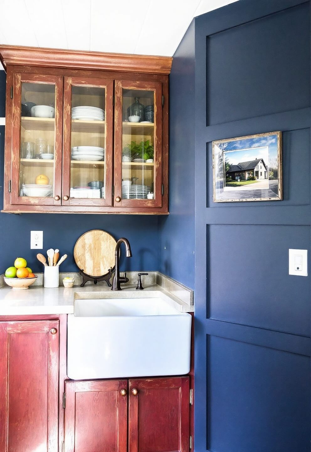 Eye-level view of a farmhouse kitchen featuring distressed red oak cabinets, glass-front uppers, antique copper hardware, deep blue walls, and a white ceramic farmhouse sink under the window