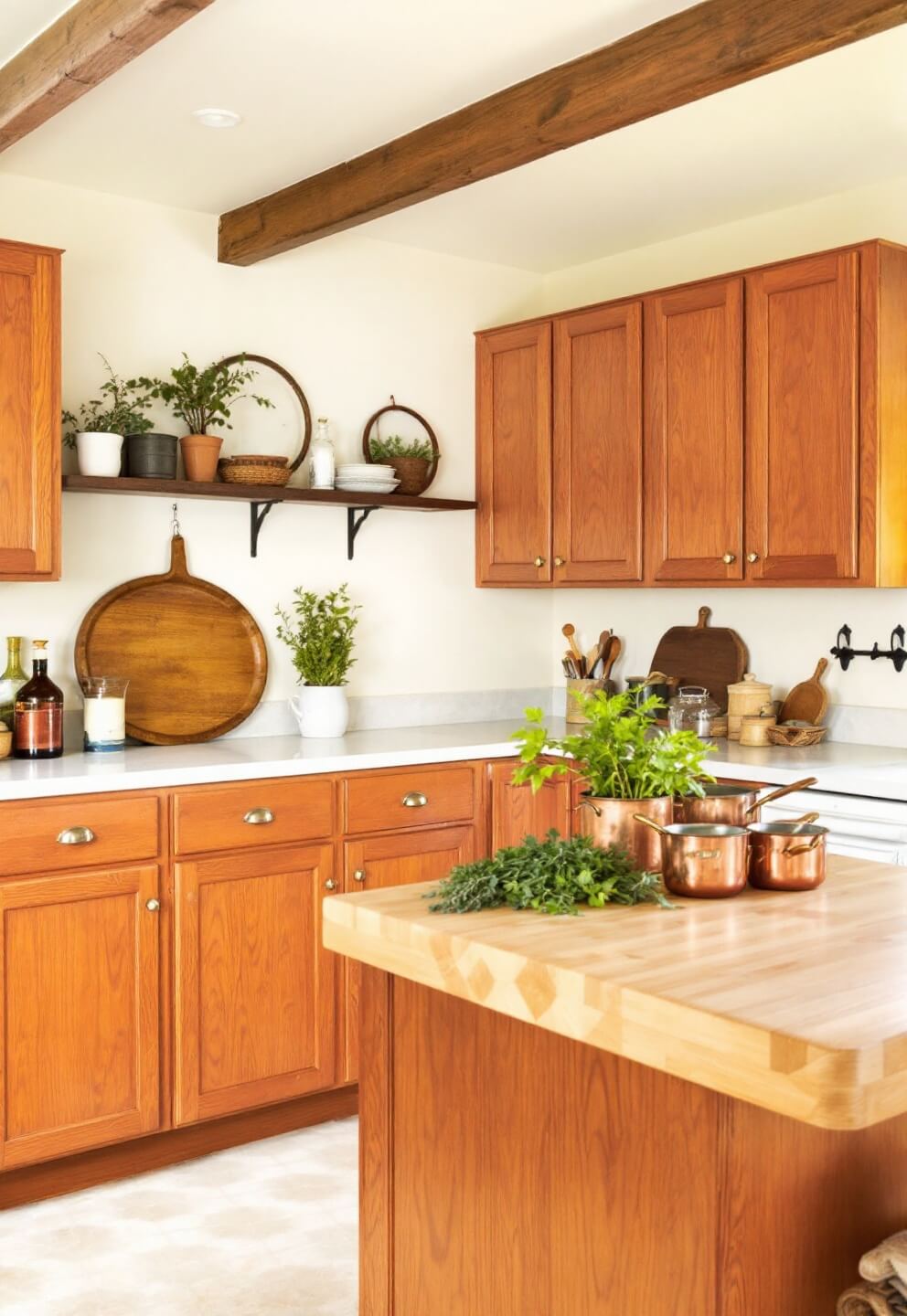 Cozy cottage kitchen with beamed ceiling, red oak cabinets, butcher block island with fresh herbs and copper cookware, cream walls adorned with vintage accessories in a corner composition for depth and warmth.