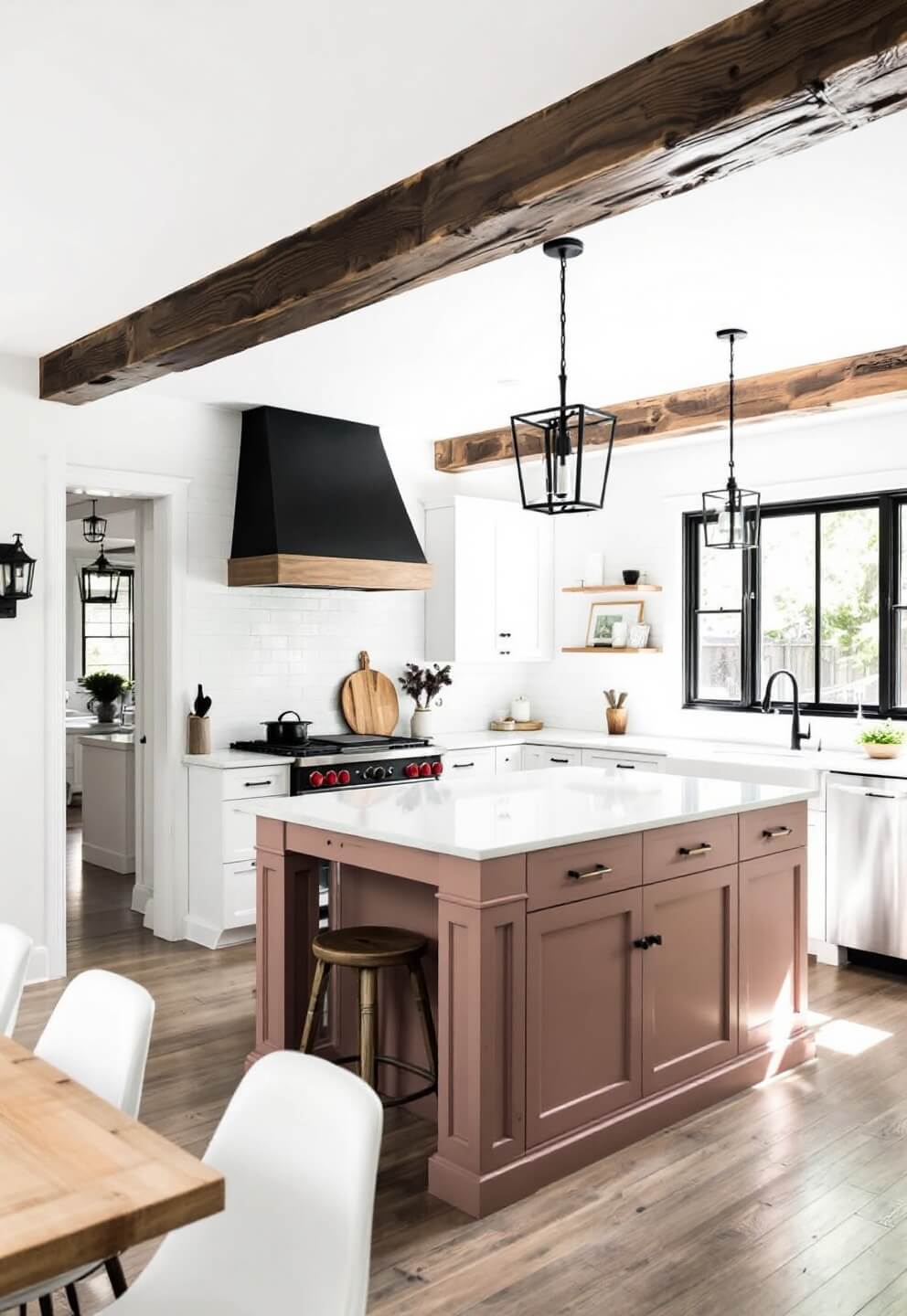 Midday shot of a modern farmhouse kitchen with dusty rose island, white cabinets, reclaimed wood beams on a 12-foot ceiling and contrasting black iron pendants and hardware, viewed from the dining area.