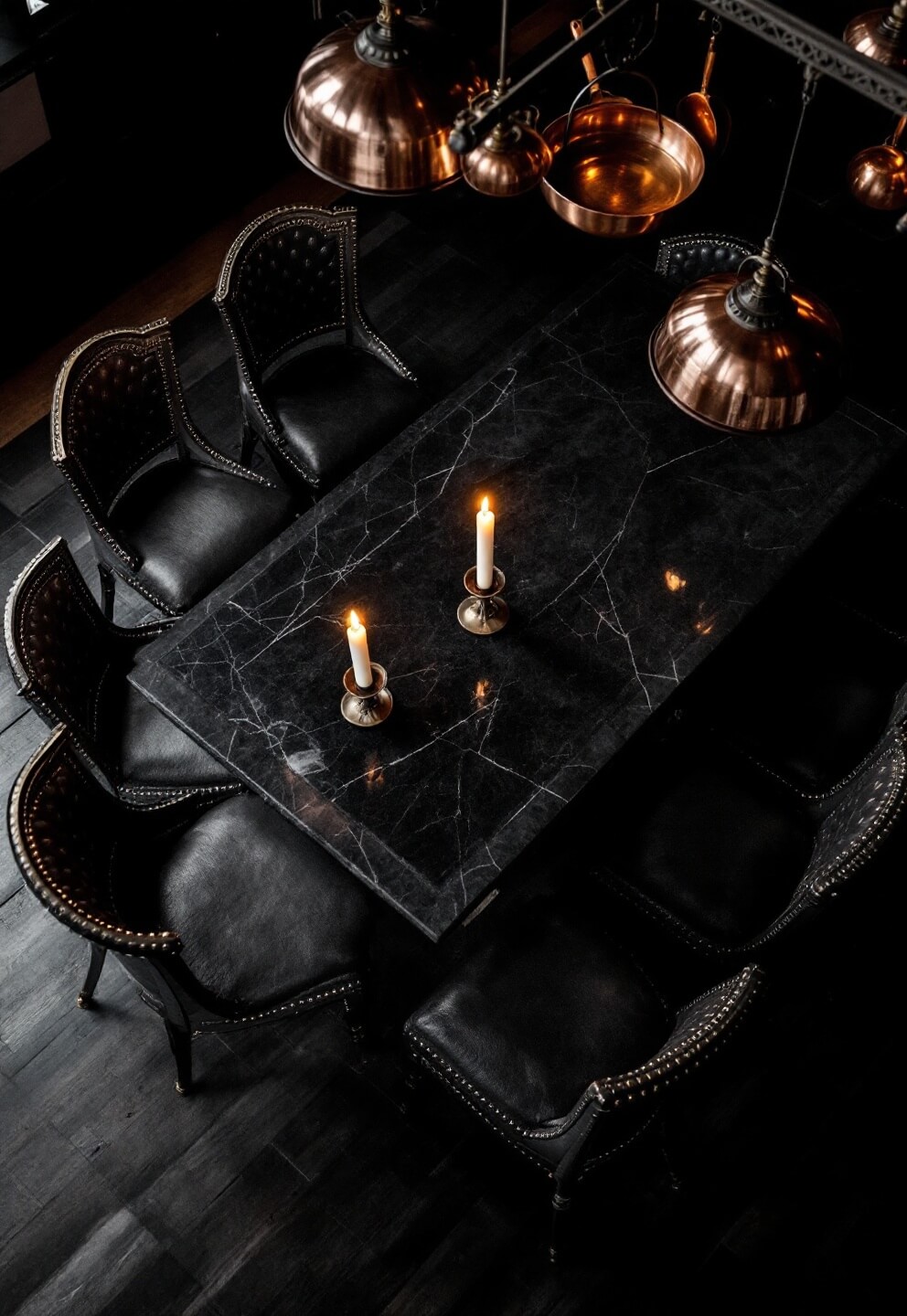 Aerial view of a Gothic-inspired Western kitchen island at night, featuring candlelit black marble surfaces, high-backed leather chairs with brass studs, Gothic pendant lights, and aged copper cookware hanging from a ceiling rack.