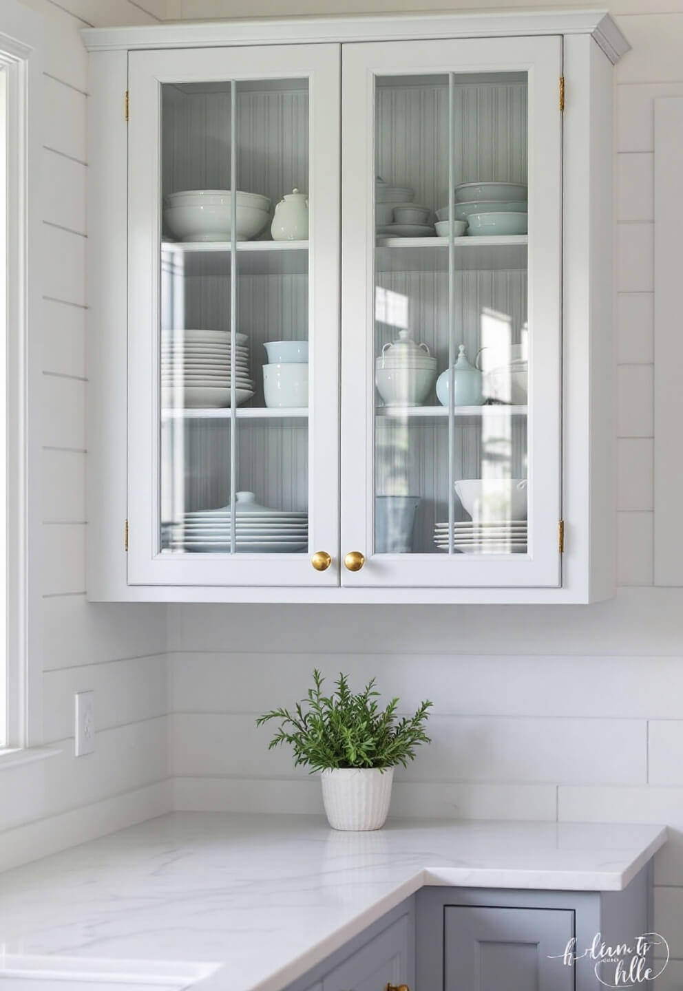 Corner detail of a coastal kitchen with a white shiplap wall and glass-front cabinet showcasing white ironstone and blue pottery under diffused morning light, with a small potted rosemary on a quartz countertop.