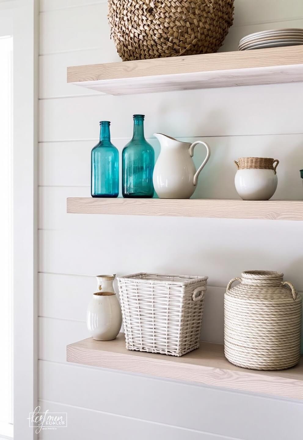 Morning light illuminating a coastal farmhouse styled open shelving with white oak shelves, vintage blue glass bottles, ceramic pitchers, woven baskets, and sea grass storage vessels against shiplap backdrop.