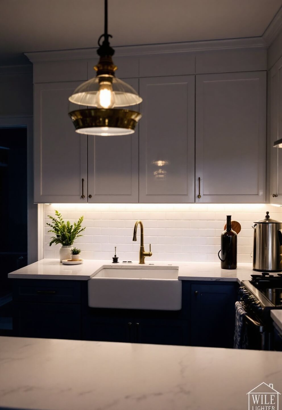 L-shaped kitchen with navy blue and white cabinets, under-cabinet LED lighting, brass pendant lamp over farmhouse sink, and white quartz countertop at dusk.