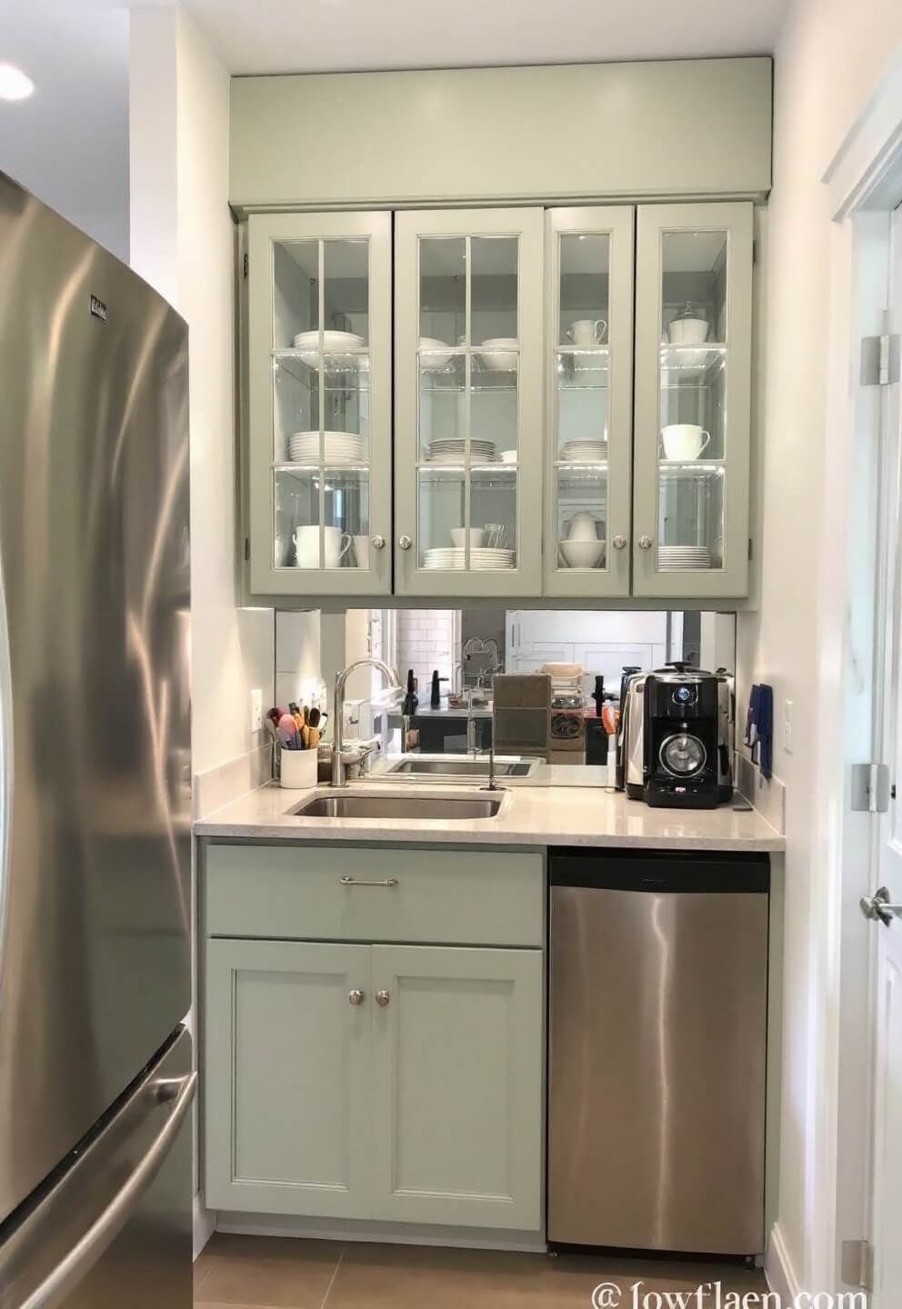 Light sage cabinets in a compact 8x10ft galley kitchen with mirrored backsplash, lit glass-fronted cabinets displaying white dinnerware, viewed through doorway for emphasis on space-saving design.