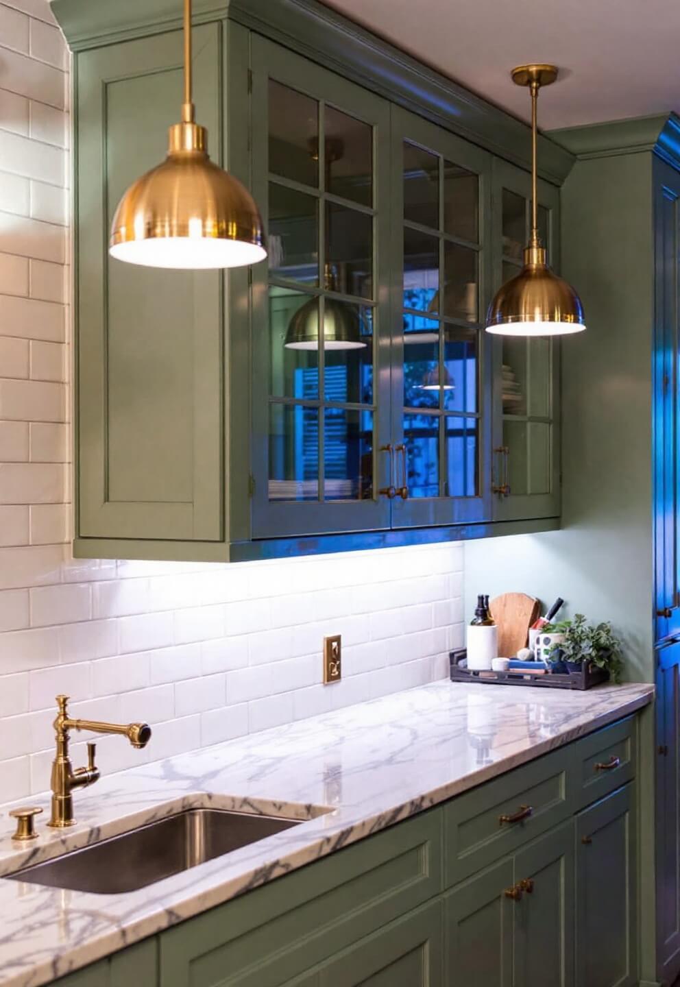 Sage green kitchen illuminated by under-cabinet LED and brass pendant lights at blue hour; light reflecting on subway tiles and marble countertop