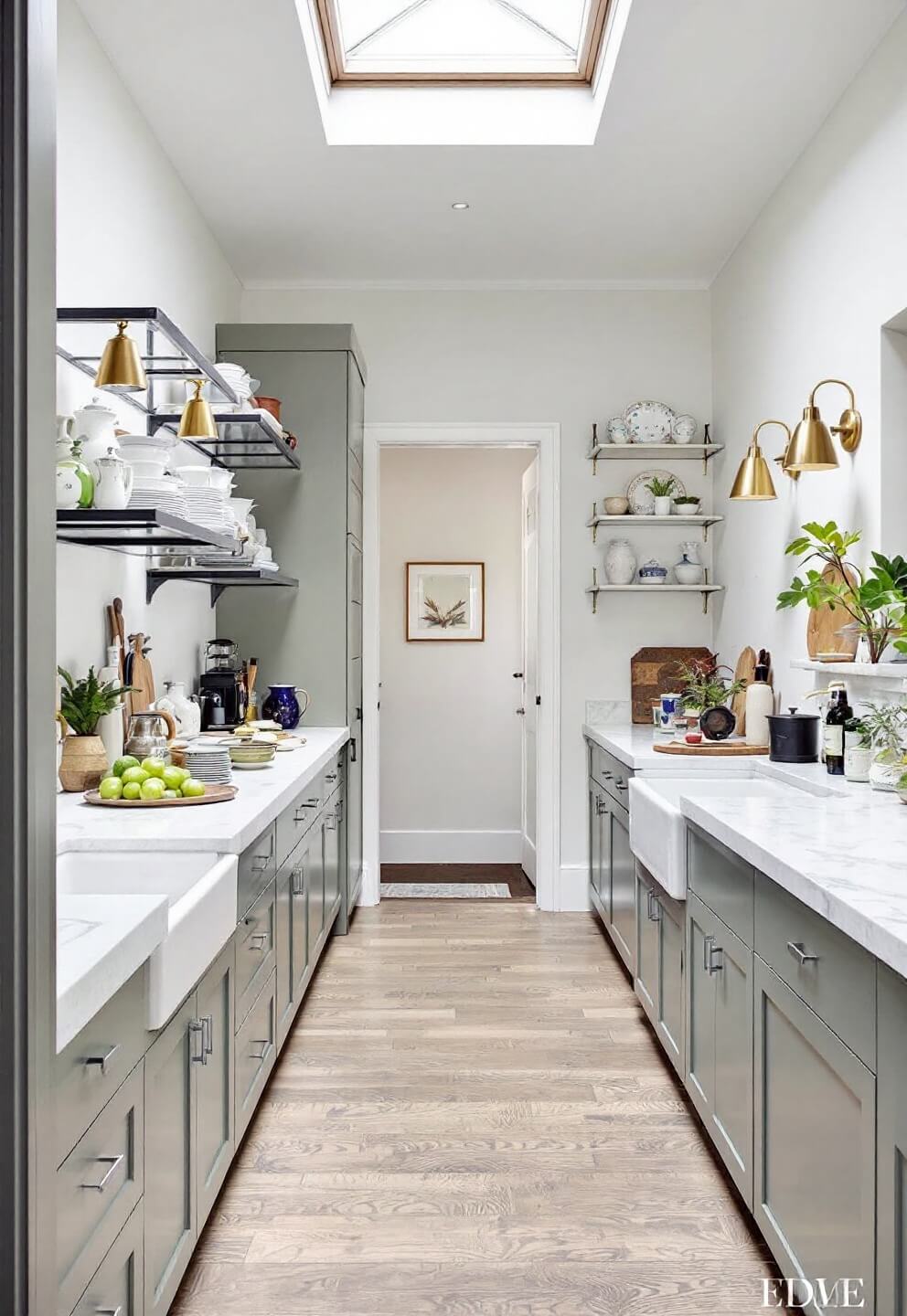 Galley kitchen with skylight, sage green cabinets, floating steel shelves with ironstone collection, soapstone counters, farmhouse sink, and brass library lights in natural light.