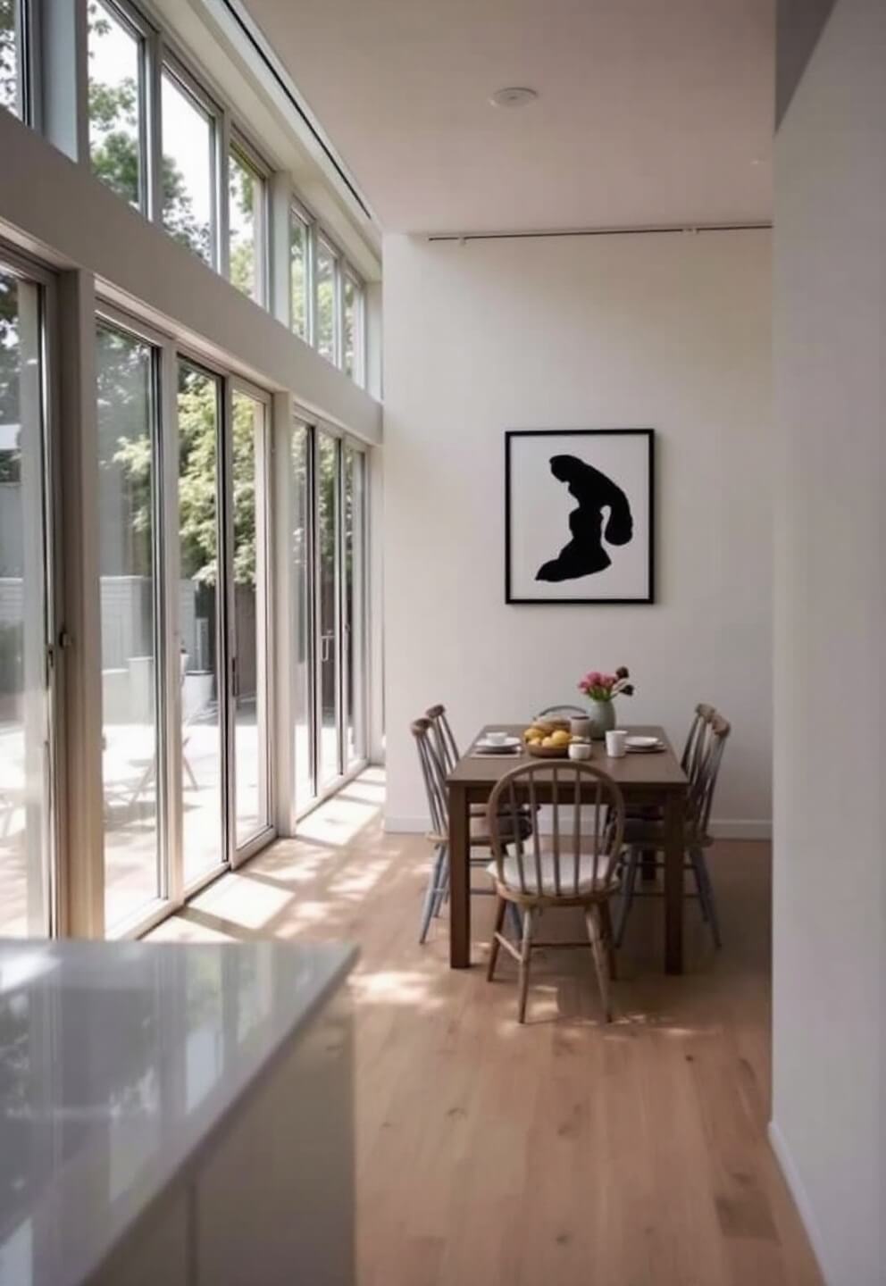 Airy kitchen-dining space with white oak flooring, floor-to-ceiling garden view windows, minimalistic black and white wall artwork, and natural light supplemented by subtle fill lighting.