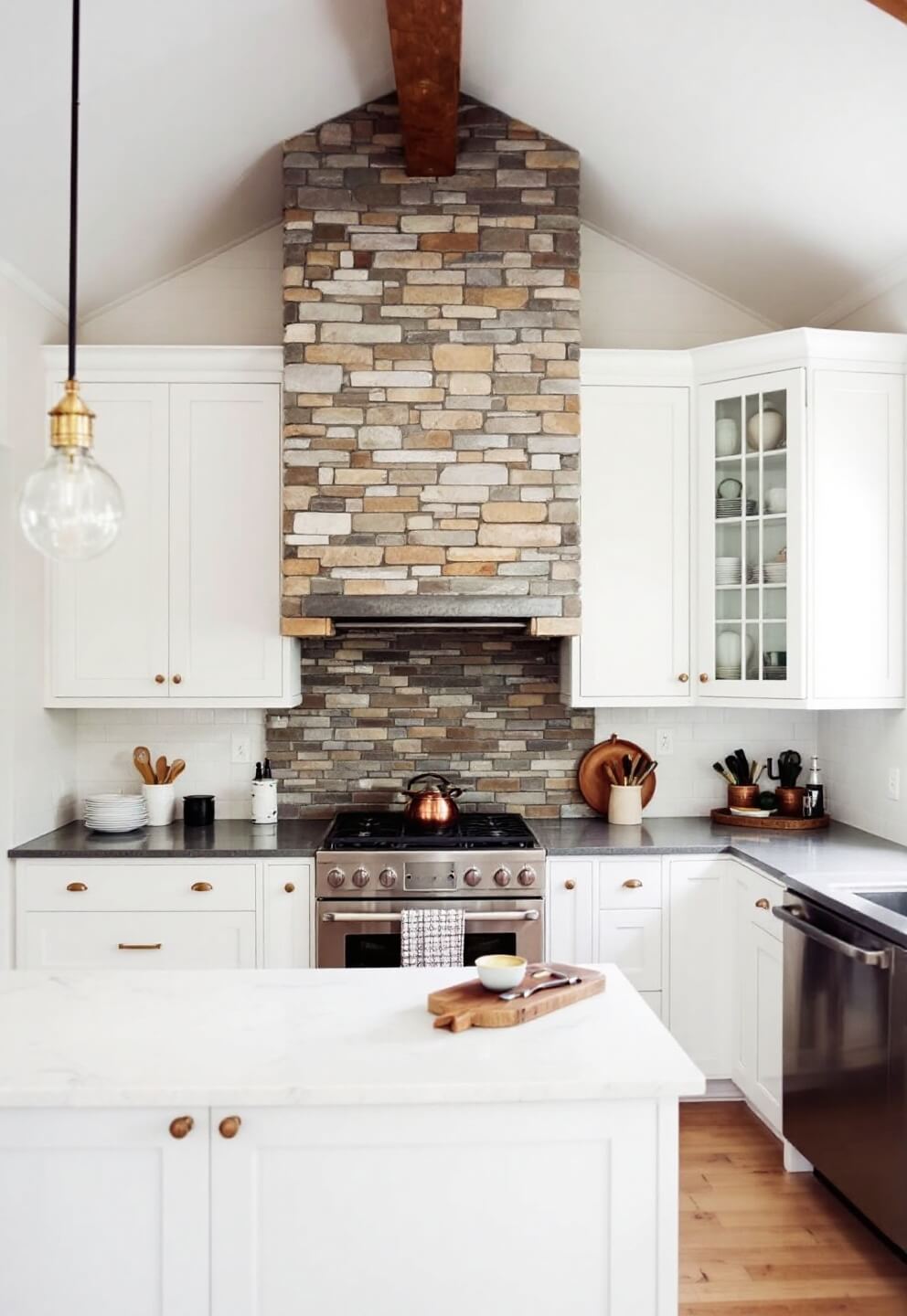 Overhead view of a spacious kitchen with vaulted ceiling, stone accent wall with custom range hood, mixed metal fixtures and a corner cabinet displaying china collection on an overcast morning.