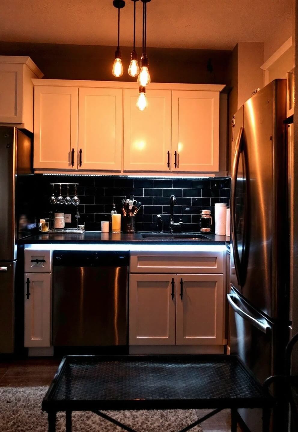 Moodily lit kitchen at night featuring under-cabinet LED lights on zinc countertops, Edison bulb pendant lights, stainless steel appliances, and a black metal bar cart with copper accents.