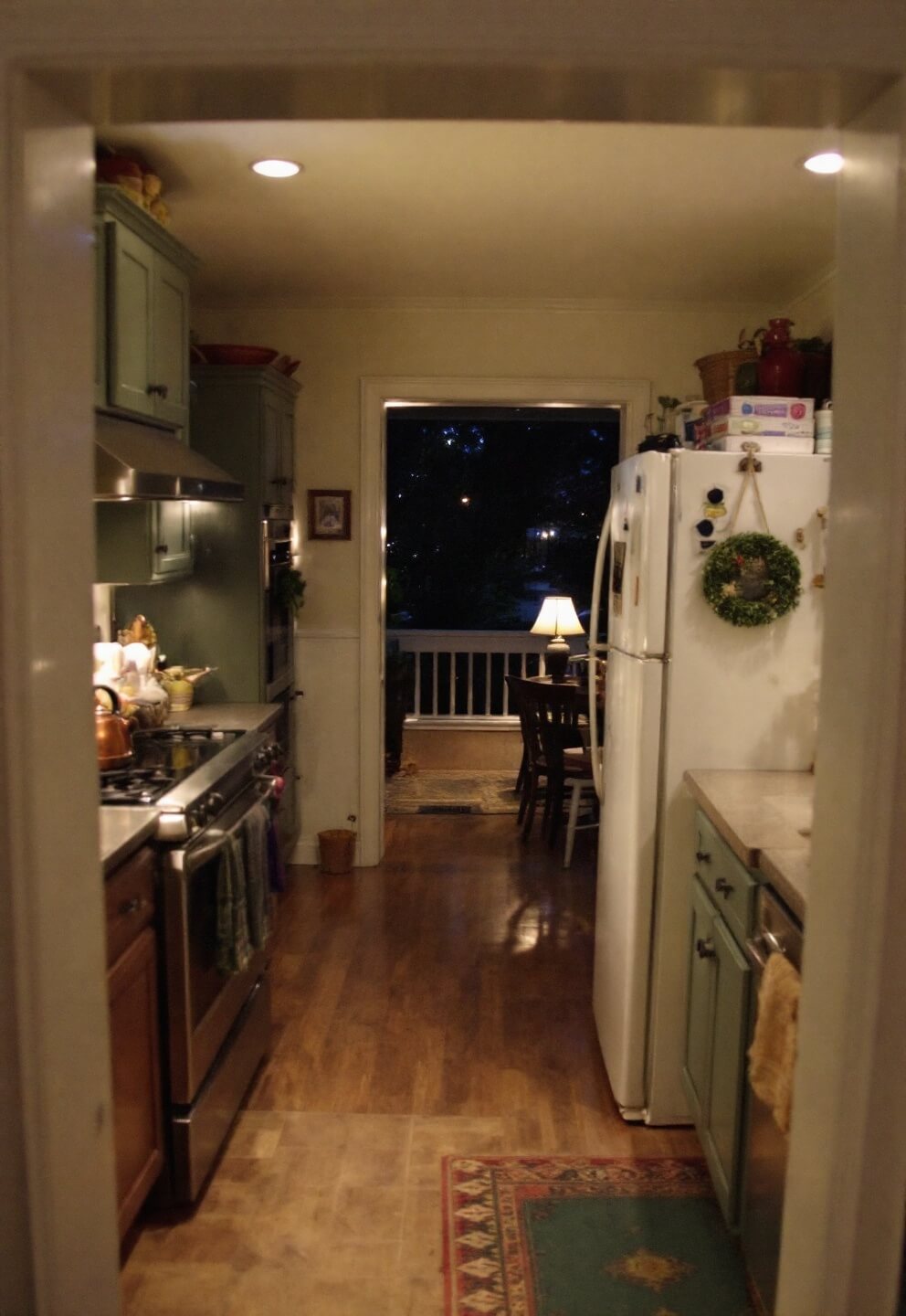 Twilight-lit kitchen in a cottage with under-cabinet lighting, soapstone counters, sage cabinetry with panel-ready fridge, and vintage-styled range with a steaming copper kettle. View from doorway highlighting depth and intimate atmosphere with multi-source lighting.