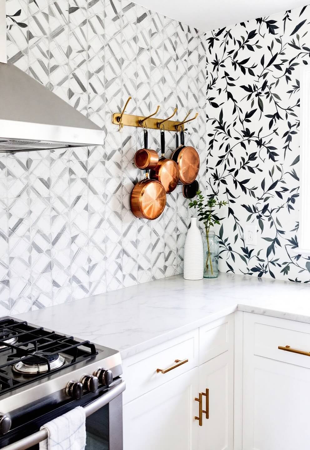 Renter-friendly kitchen makeover featuring gray and white peel-and-stick geometric backsplash, displayed copper cookware on brass hooks, marble-pattern contact paper on white cabinets, botanical removable wallpaper on one wall, and natural light illuminating the room.