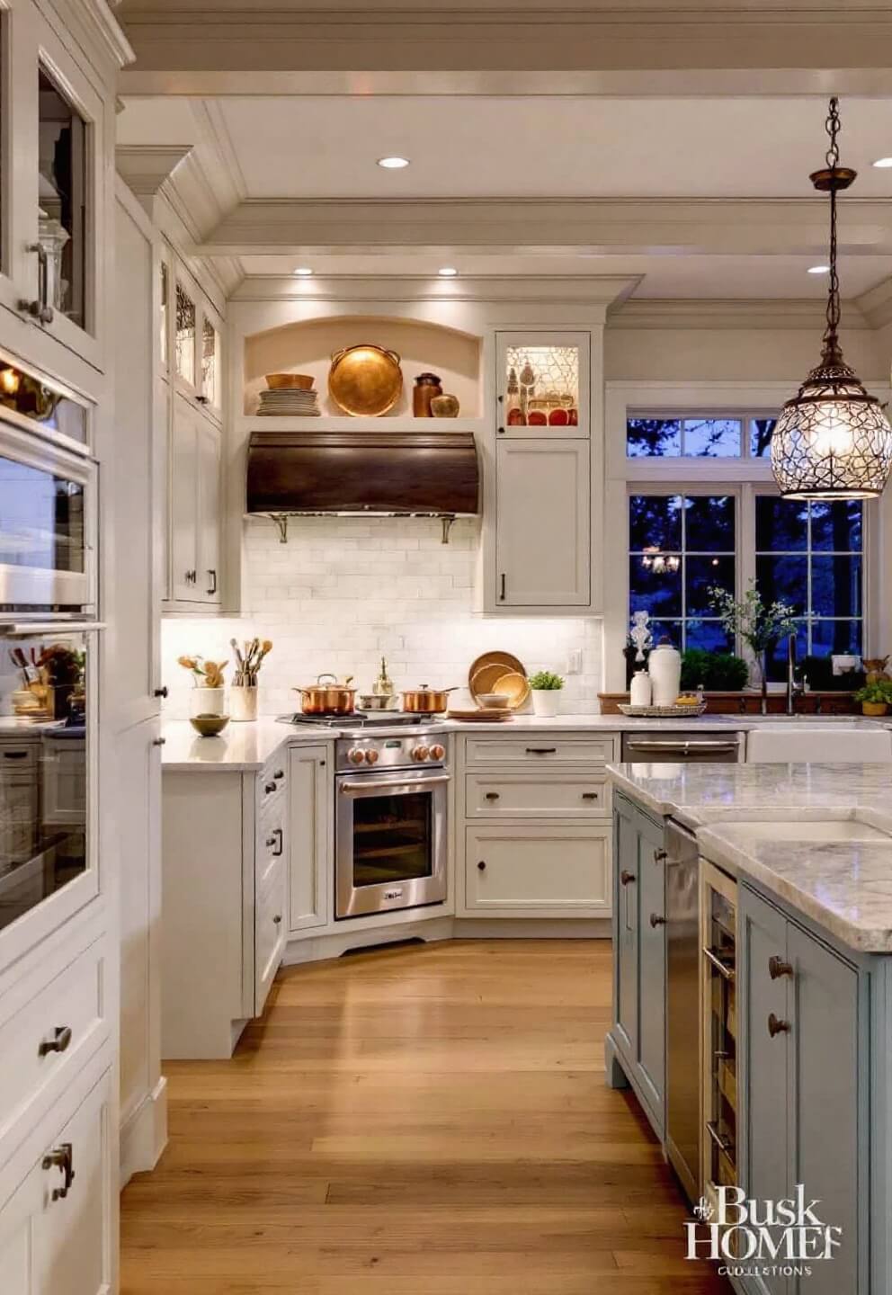 Evening view of a country kitchen with cream Shaker-style cabinets, dusty blue double-width island, open shelves with copper cookware and white ironstone and pendant lights illuminating the room