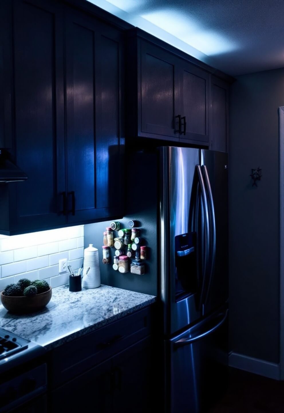 High-angle view of an organized L-shaped kitchen (12'x8') at dusk with LED strip light under cabinets, magnetic spice jars on stainless steel fridge, and white subway tile backsplash.