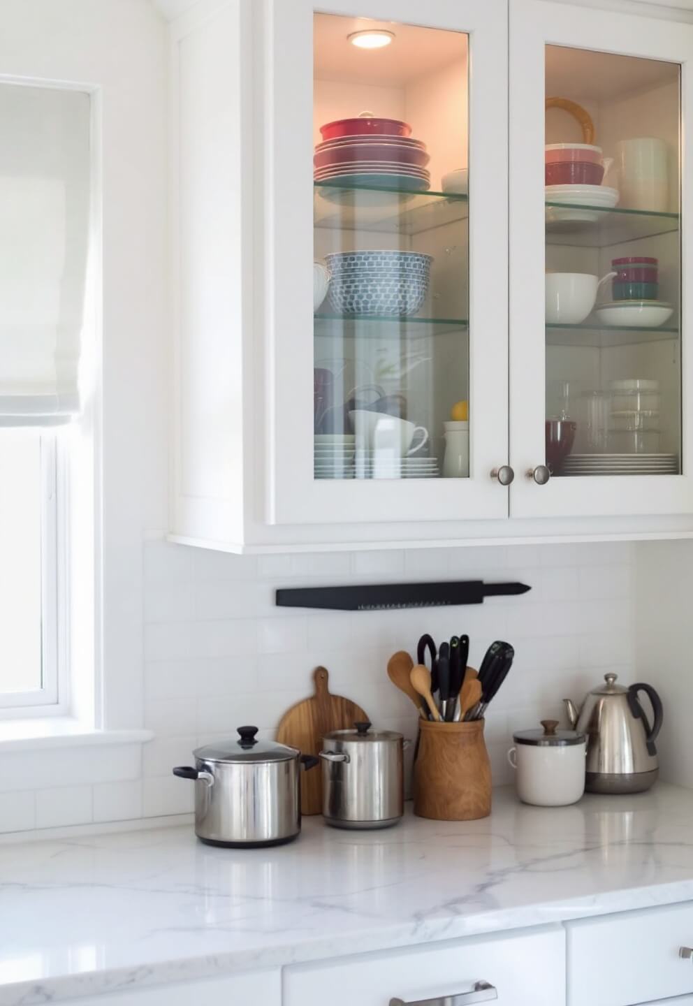 Compact kitchen with glass-front illuminated upper cabinets displaying colorful cookware, white quartz countertops, and a magnetic knife strip, with a focus on vertical storage solutions