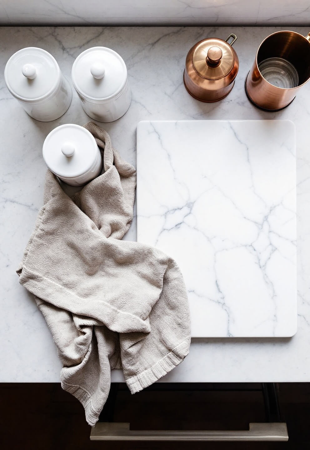 Overhead view of soapstone counter prep area with marble pastry board, vintage ceramic canisters, copper measuring cups and textured linen napkin under soft, diffused lighting