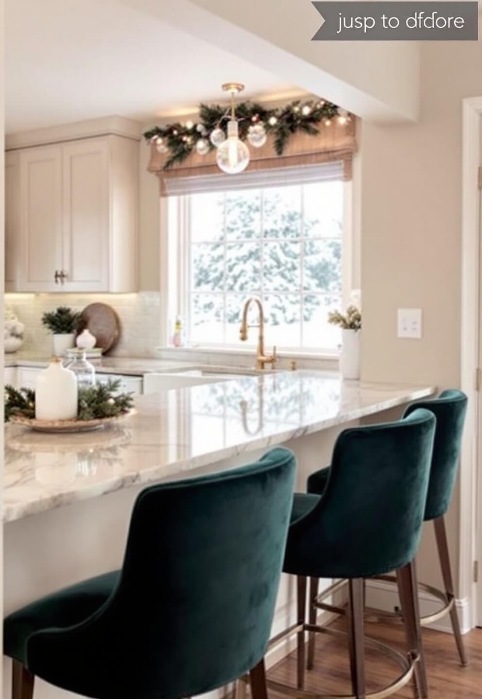 Winter morning in a kitchen with snow visible through windows, cream marble counter topped with holiday decorations and three forest green velvet barstools, warmed by under-cabinet lighting.