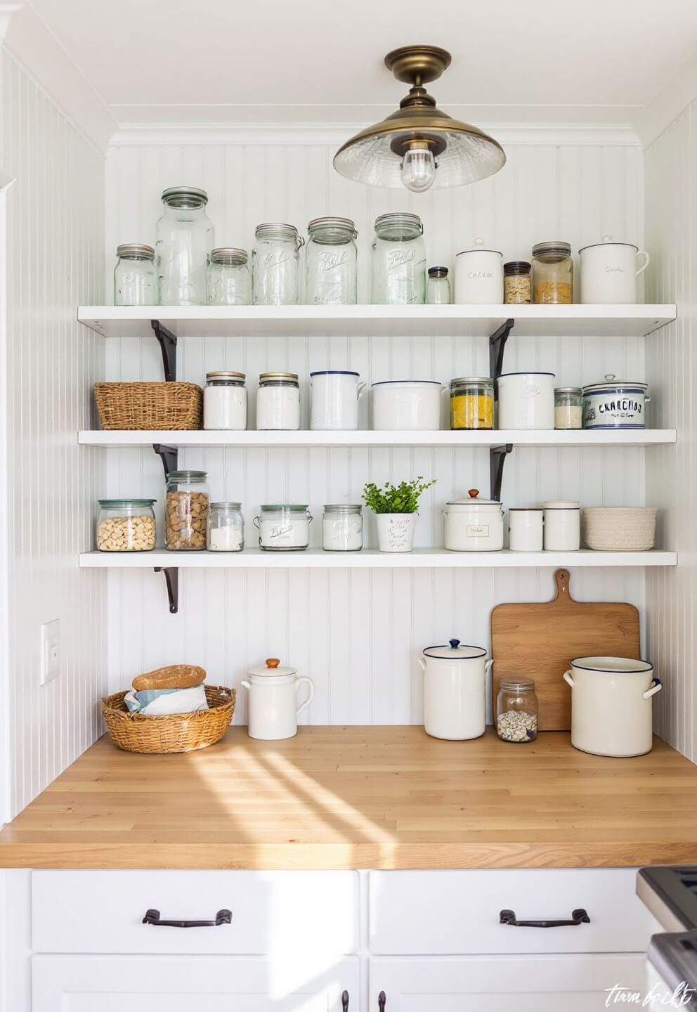 Morning light illuminating an organized 8x8 ft kitchen pantry with open shelving displaying mason jars, vintage canisters, enamelware, and a butcher block counter with bread board and ceramic crock on beadboard walls in warm white.