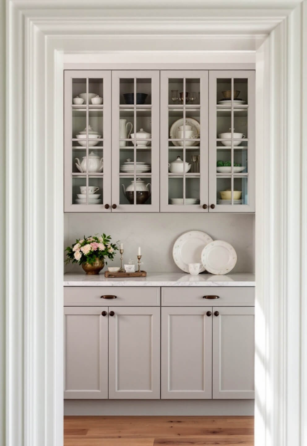 Butler's pantry with dove grey wood cabinetry, glass-fronted cabinets displaying heritage china, marble countertop with antique silver and crystal, captured in afternoon light through doorway frame