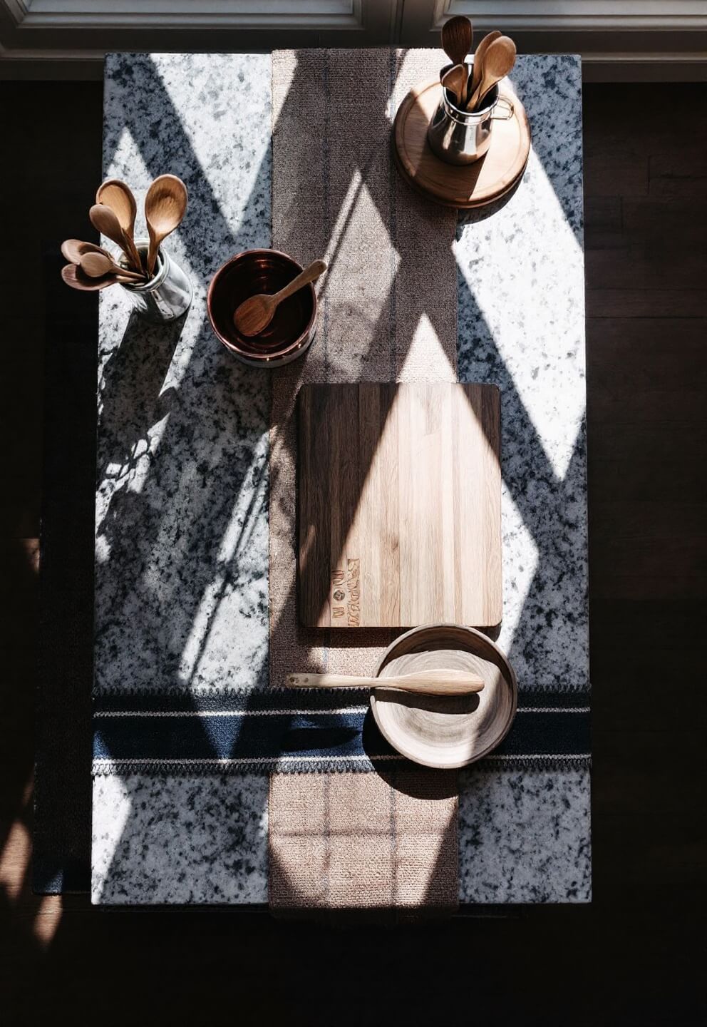 Overhead drone shot of a well-organized kitchen island workspace with textured granite countertop, inlaid butcher block, navy and terra cotta woven runner, copper bowls and wooden utensils under natural skylight illumination.