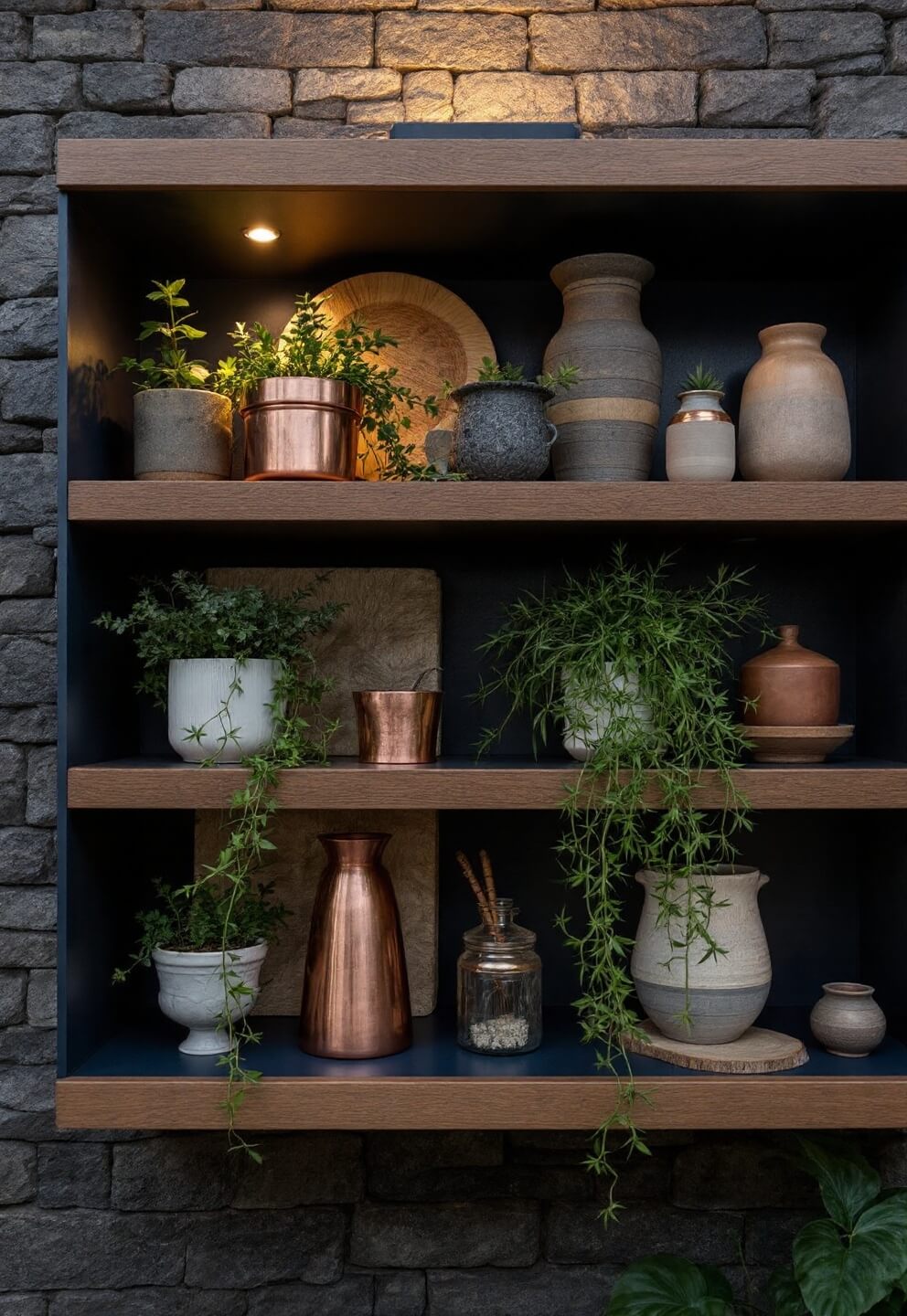 Close-up of copper, wood, and ceramic collectibles on navy painted shelves against a textured stone wall, illuminated by warm LED lights at twilight with trailing plants adding an organic touch.