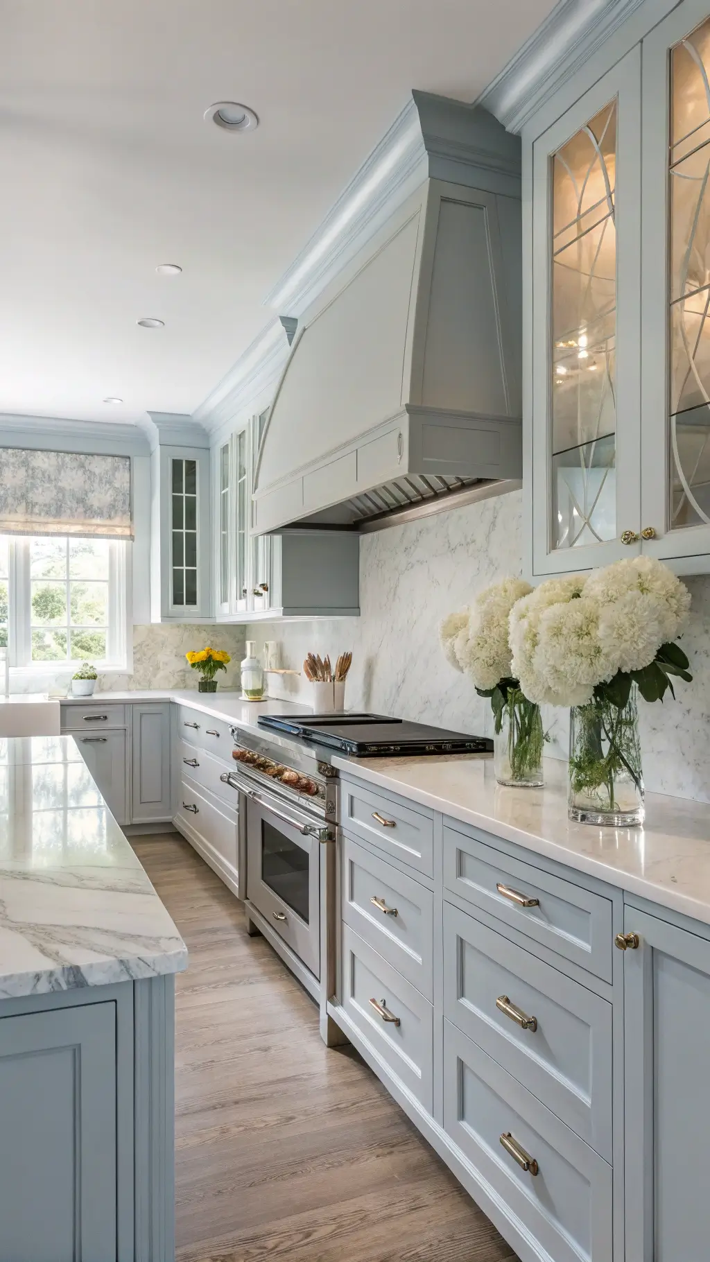 Elegant transitional kitchen with powder blue inset cabinets, quartzite counters, custom range hood, crystal knobs, and white hydrangeas in mercury glass vases, bathed in soft afternoon light through roman shades