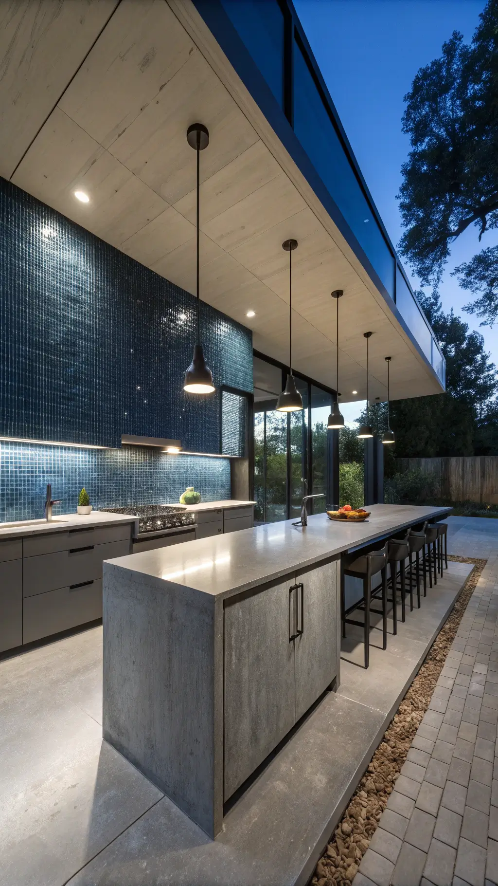 Open-concept contemporary kitchen at dusk with midnight blue glass penny tile backsplash, grey cabinets, concrete counters, black matte fixtures, and dramatic shadow-casting pendant lights.